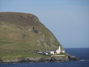 Lighthouse on a rocky cliff, surrounded by green meadows and blue sea under a clear sky, white