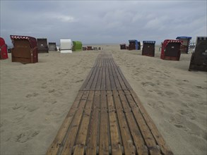 A wooden walkway leads through the sand, lined with beach chairs under a cloudy sky, beach chairs