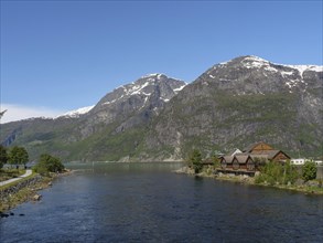 A calm river with houses on its banks, surrounded by snow-capped mountains and forest under a clear