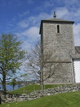 Stone church tower standing in a green lawn with trees and blue sky, old stone church and many