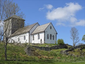 White church with stone tower on a hill under a blue sky, trees in the foreground, old stone church