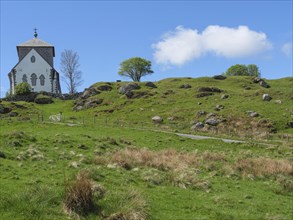 Church with stone tower on hilly landscape under blue sky with clouds, trees in the background, old