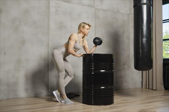Female bodybuilder pushing dumbbell in the gym leaning on metal barrel