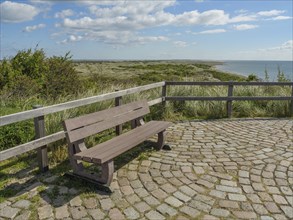 Bench on a paved floor with a view of the sea and the dunes, under a blue sky with clouds, dune and