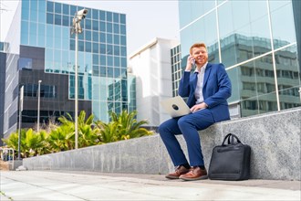 Businessman talking to the mobile while working with laptop sitting outside an office block in the