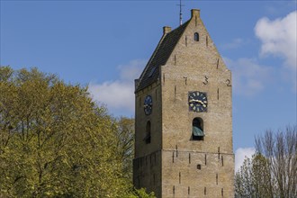 A medieval bell tower with clocks on both sides against a blue sky, historic houses and a church