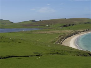 The natural backdrop shows a sandy coastline surrounded by green meadows and hills, complemented by