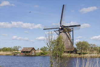 A windmill and a shed next to a body of water, surrounded by reeds under a sunny sky, many historic