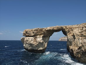 Large rock arch towers over the sea, surrounded by cliffs and under a clear sky, many historic