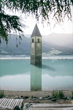 Sunken tower in Lake Reschen with mountains in the background and a gravel path in the foreground.