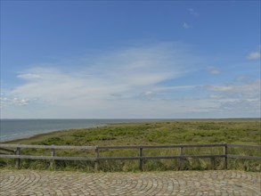 Panoramic view of the dunes and the sea behind a wooden fence and a paved floor under a blue sky,