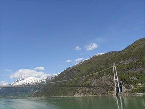 Wide view of a bridge and snow-covered mountains over a green lake and clear blue sky, bridge in a