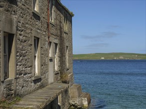 Empty stone building with several windows on the edge of clear blue water and a clear sky in the