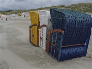 Three colourful beach chairs next to each other on a sandy beach under a cloudy sky, colourful