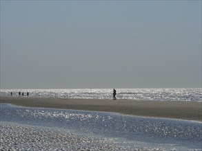 Single person walking along a wide beach, other people visible in the distance, glittering sea