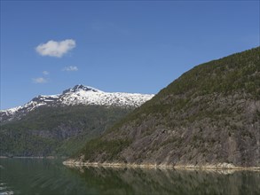 A calm lake surrounded by high green mountains with snow-capped peaks under a clear blue sky, calm