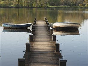 Wooden jetty leading into the water with two rowing boats on a quiet lake surrounded by trees,