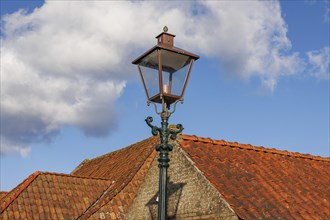 Classic street lamp in front of a tiled roof and blue sky with single clouds, old houses and small
