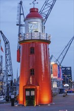 A striking red lighthouse illuminated at night, surrounded by harbour cranes and buildings, small