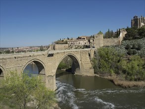 Stone bridge over a river with historic buildings and vegetation under blue sky, historic stone