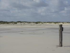 A wooden beam stands alone on a white sandy beach with dunes in the background under a cloudy sky,