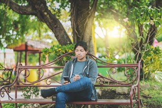 Handsome man sitting on a bench checking cell phone at sunset. Young man sitting on a park bench