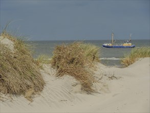 Sand dunes with grass in the foreground, a blue ship on the sea and a cloudy sky, dunes on an