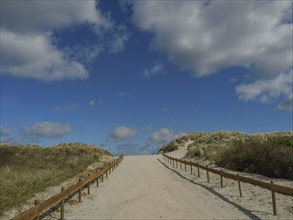 A wide sandy path leads through the dunes, surrounded by a clear blue sky and clouds, dunes by the