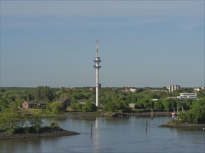 A television tower surrounded by green trees and houses on a quiet river under a clear blue sky,