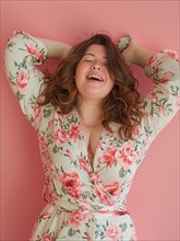 Excited curvy caucasian woman in a floral dress laughing with her hands raised, against a pink