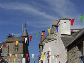 Street scene with colourful pennants hanging between buildings and a tower, calm sea with rocks and