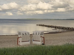 Two white beach chairs on a sandy beach, rocks in the water and cloudy sky in the background, beach