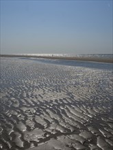 Wide beach with undulating sand surface and reflecting watercourse under a clear sky, glittering
