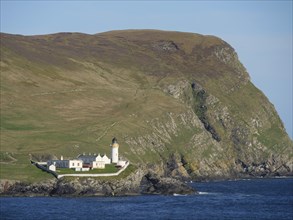 A lighthouse and some houses stand on cliffs on the coast, surrounded by grassy hills and under a