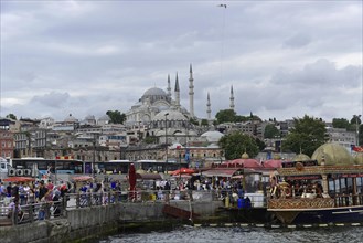 A busy harbour market with boats in the foreground. A mosque and minarets can be seen in the