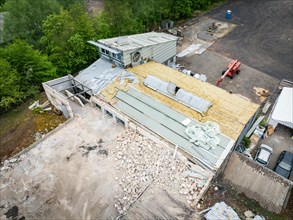 A construction site with a partially demolished building, containers and surrounded by green trees