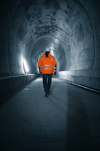 A person in a safety waistcoat walks through an illuminated tunnel with concrete walls, which has a