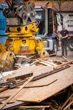 An excavator piles wood debris next to a lorry while a worker stands nearby, demolition,