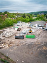 A construction site with containers and rubble, surrounded by trees and hills, with houses in the