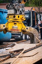 An excavator with a hydraulic grab stacks wood debris while a worker stands nearby, demolition,