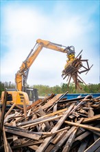 Large excavator lifts wood debris on a construction site under a cloudy sky and numerous