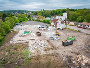 A construction site with demolished material, containers and machines, surrounded by green trees