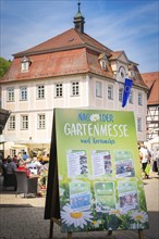 A poster for a garden fair is shown in front of a historic building in sunny weather, spring,
