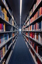 A narrow corridor between high bookshelves in a library with multi-coloured books that radiate a