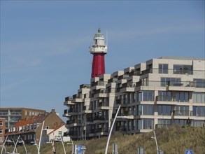 Modern lighthouse next to residential buildings under a clear blue sky, parasols and a pier on the