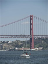 Sailboat in front of a large red bridge over a wide river in an urban environment, Lisbon,
