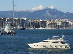 Harbour with a yacht in the foreground, city and mountains in the background under a cloudy blue