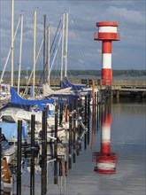 A harbour with sailing boats and a red and white lighthouse reflected in the calm water, cloudy