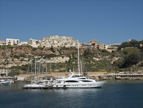 Yacht and boats in the harbour next to a hilly coastal town, the island of Gozo with historic