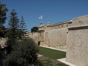 Historic fortress with protective stone walls, wide green spaces and a waving flag under a bright